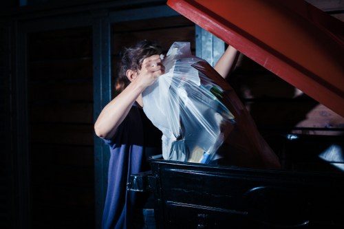 A person with short, dark hair is shown disposing of large plastic garbage bags filled with mixed rubbish into a black wheelie bin inside a dimly lit storage area or garage. The individual is wearing a dark T-shirt and is holding a clear plastic bag, which is partially visible, containing various waste items. The scene reveals a wooden wall in the background with visible horizontal planks and a metal framework, suggesting an urban or residential environment typical of properties in Notting Hill. The plastic bags appear to be filled with general waste, possibly from household or business clearance, and are being carefully placed into the bin in preparation for rubbish removal. The lighting highlights the person's face and the reflective surfaces of the plastic bags, emphasizing their focus on the task. This image exemplifies aspects of waste disposal services offered by Commercial Waste Removal Notting Hill, including handling and correctly disposing of waste materials in a secure and organized manner within a typical indoor or semi-outdoor space near central London postcodes.