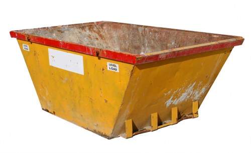 A large, industrial skip bin painted in bright yellow with a red trim along the top edge, situated outdoors on a paved surface. The bin's interior is visible and appears to be rusted, with a textured, weathered metallic finish. The sides of the skip are slightly stained and marked, indicating previous use for waste collection. The front of the bin shows small, yellow support brackets or feet at the base, and the overall shape is rectangular with a wide open top for easy loading. The outdoor environment in the background is plain, with no other objects or structures visible, suggesting it may be part of a commercial waste removal operation in a business or industrial area, likely near Notting Hill. The image emphasizes the functionality of the skip for rubbish collection and waste removal services provided by Commercial Waste Removal Notting Hill, aligning with their business clearance and rubbish disposal services. The lighting is natural, enhancing the vividness of the yellow and red colours, and the focus is sharp on the bin's surface details.