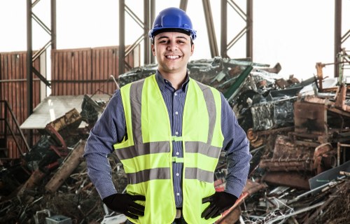 A young male waste management worker indoors at a scrapyard or waste storage facility, wearing a blue safety helmet, a high-visibility yellow vest over a dark shirt, and black gloves. He is standing with his hands on his hips, smiling confidently towards the camera. Behind him, there is a large pile of assorted scrap metal, including car parts and other metallic debris, some rusted and weathered. The interior features metal framing and natural light filtering through the structure, indicating a spacious industrial environment typical of waste collection and disposal sites in Notting Hill or nearby areas. The scene demonstrates a professional approach to rubbish and waste management services, with a focus on handling mixed metallic waste materials.
