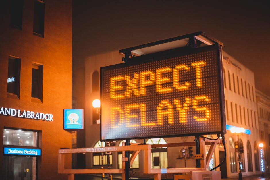 A large, illuminated digital warning sign mounted on a metal frame displays the message 'EXPECT DELAYS' in bright orange LED lights. The sign is positioned on a city street at night, with the backgrou